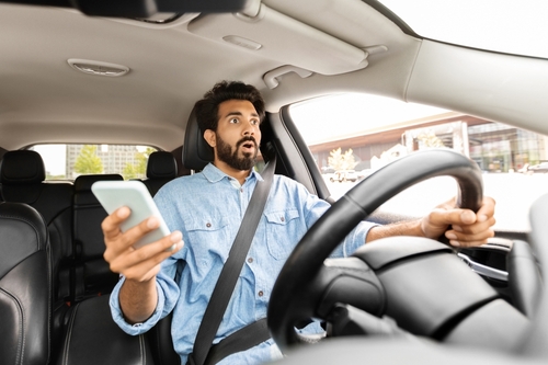 Man driving a car, looking shocked while holding a smartphone, illustrating the dangers of distracted driving in rideshare situations.