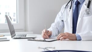 Doctor in a white coat writing notes on a clipboard with a laptop nearby, emphasizing the importance of medical records in car accident cases.
