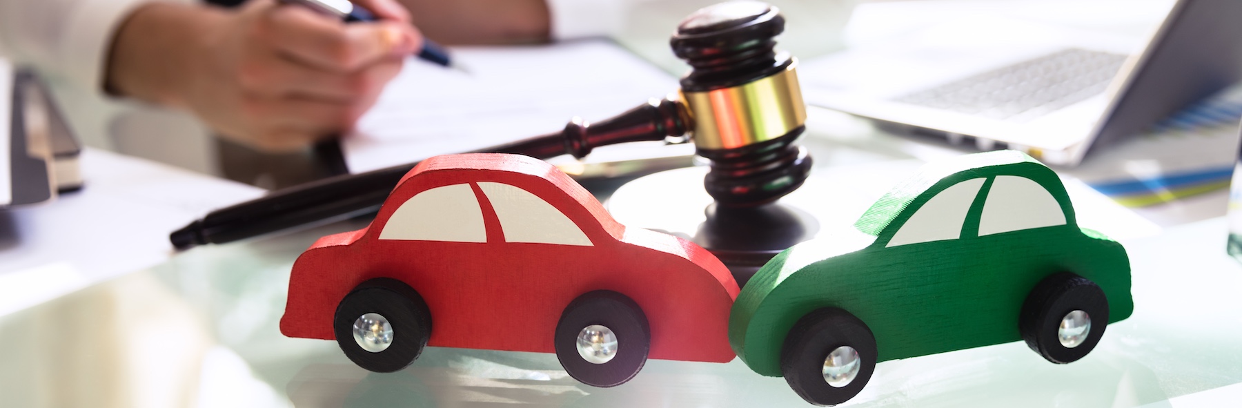 Red and green wooden toy cars positioned in front of a judge's gavel on a glass desk while a person writes legal documents, representing car accident attorneys at work.