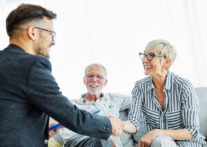 A young male attorney in a dark blazer and glasses, shaking hands with a smiling senior woman in striped shirt while a senior man watches providing local legal guidance consultation.