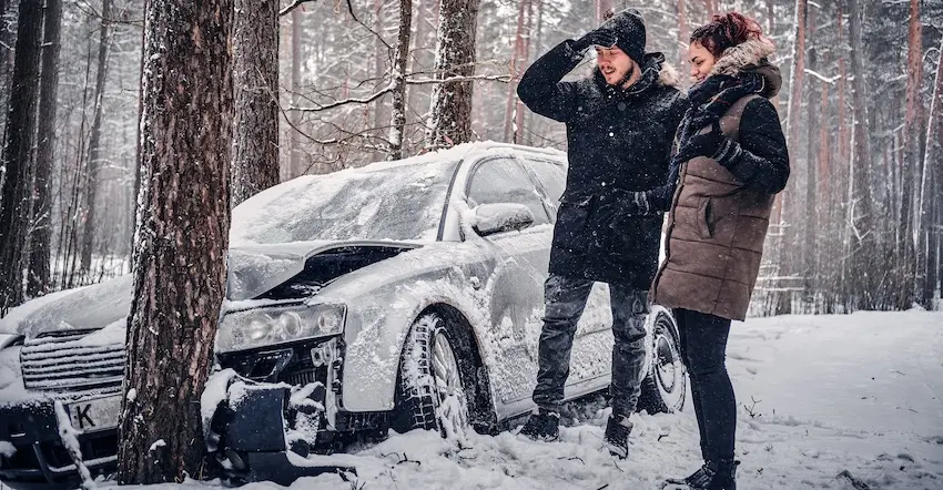A man and woman in winter coats stand beside a snow-covered car that has crashed into a tree in a snowy forest depicting the dangerous road conditions that contribute to car accidents during winter storms in North Carolina.