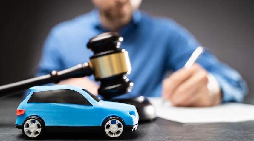 A blue toy car sits in the foreground beside a judge's gavel while a man in a blue shirt signs a document in the background representing the legal settlement process with North Carolina Car Accident Lawyers that resolves common car accident claims through negotiation or court proceedings in North Carolina.