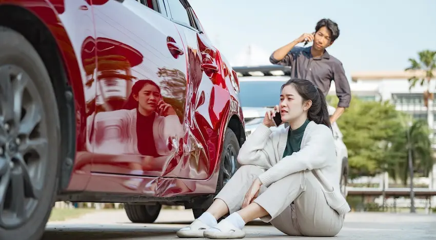 A distressed young woman sits on the ground against a damaged red car while talking on her phone and a man makes a call in the background depicting the immediate aftermath scene that gives rise to some of the most common car accident claims filed each year in North Carolina.