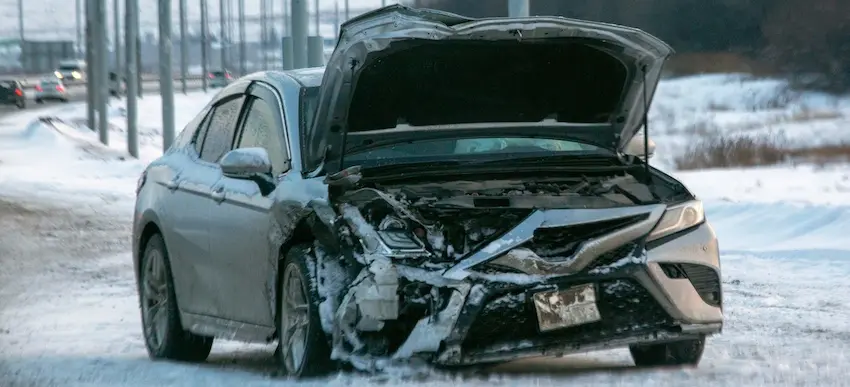 A severely damaged silver sedan sits on a snow-covered road with its hood crumpled and trunk popped open following a winter collision showing the extent of vehicle destruction that supports common car accident claims for property damage and personal injury with help from North Carolina Car Accident Lawyers.