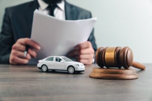 An attorney in a dark charcoal suit and black tie sits at a dark wood desk in the blurred background holding a multi-page white legal document open in both hands and reviewing it carefully while in the sharp foreground a detailed white toy sedan sits on the desk surface beside a polished honey-toned wooden judge's gavel with a gold band resting upright on its matching circular sound block against a clean light gray background — a legal concept image representing the case review and documentation work a car accident lawyer from North Carolina Car Accident Lawyers performs when building a client's claim.
