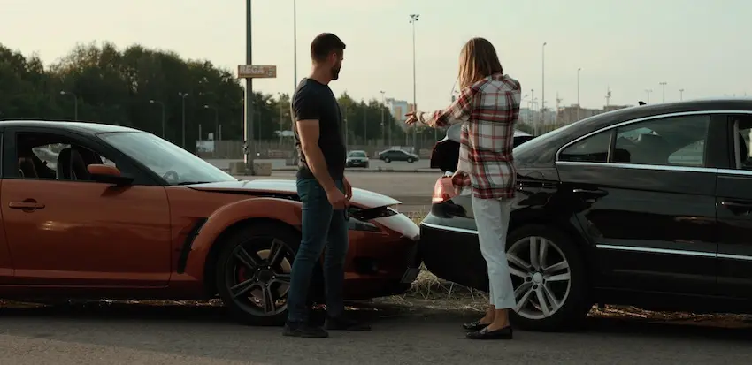 A man in a dark navy t-shirt and jeans stands with his hands at his sides facing a woman in a red and white plaid flannel shirt and white jeans who is gesturing expressively with one hand while holding a smartphone in the other- both standing between two vehicles that have been involved in a rear-end collision — a bright orange muscle car on the left with front-end damage and a black sedan on the right with its rear bumper crumpled — in an open parking area with stadium light poles- trees- and a blurred urban skyline visible in the fading evening light behind them- depicting the tense post-collision exchange between two drivers before each contacts a car accident lawyer in North Carolina.