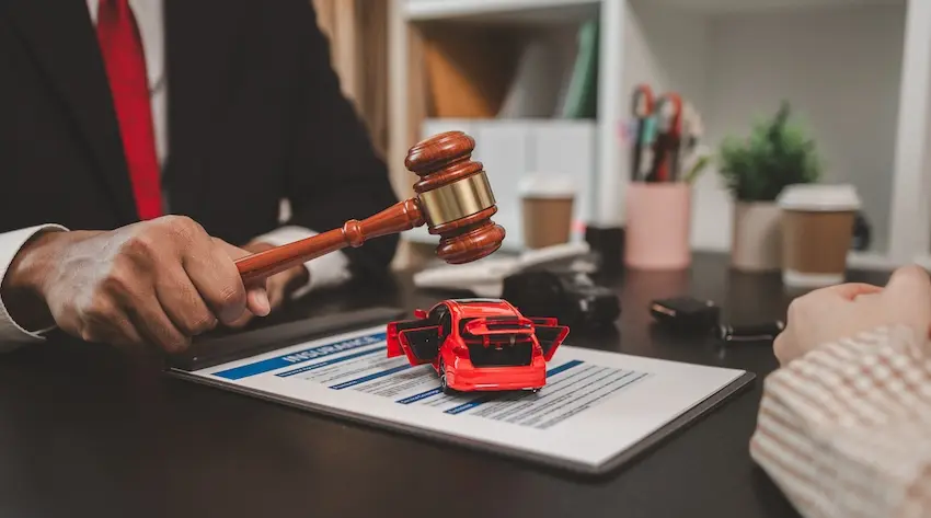 A man in a dark suit with a red tie extends one hand holding a wooden judge's gavel with a gold band downward over a dark desk surface where a red toy sedan with its doors open sits positioned on top of a printed insurance document on a clipboard while another person's hand in a striped sleeve is visible on the right side of the frame with a blurred office background showing bookshelves- a small potted plant- a coffee cup and a set of car keys on the desk — illustrating the formal legal and insurance claim process overseen by a car accident lawyer- from North Carolina Car Accident Lawyers in North Carolina- on behalf of an injured client.